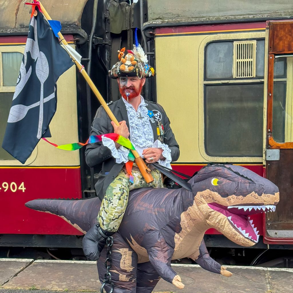 Person in a colorful pirate-like costume with a dragon inflatable prop tied to their legs, standing beside a train car and carrying a flag