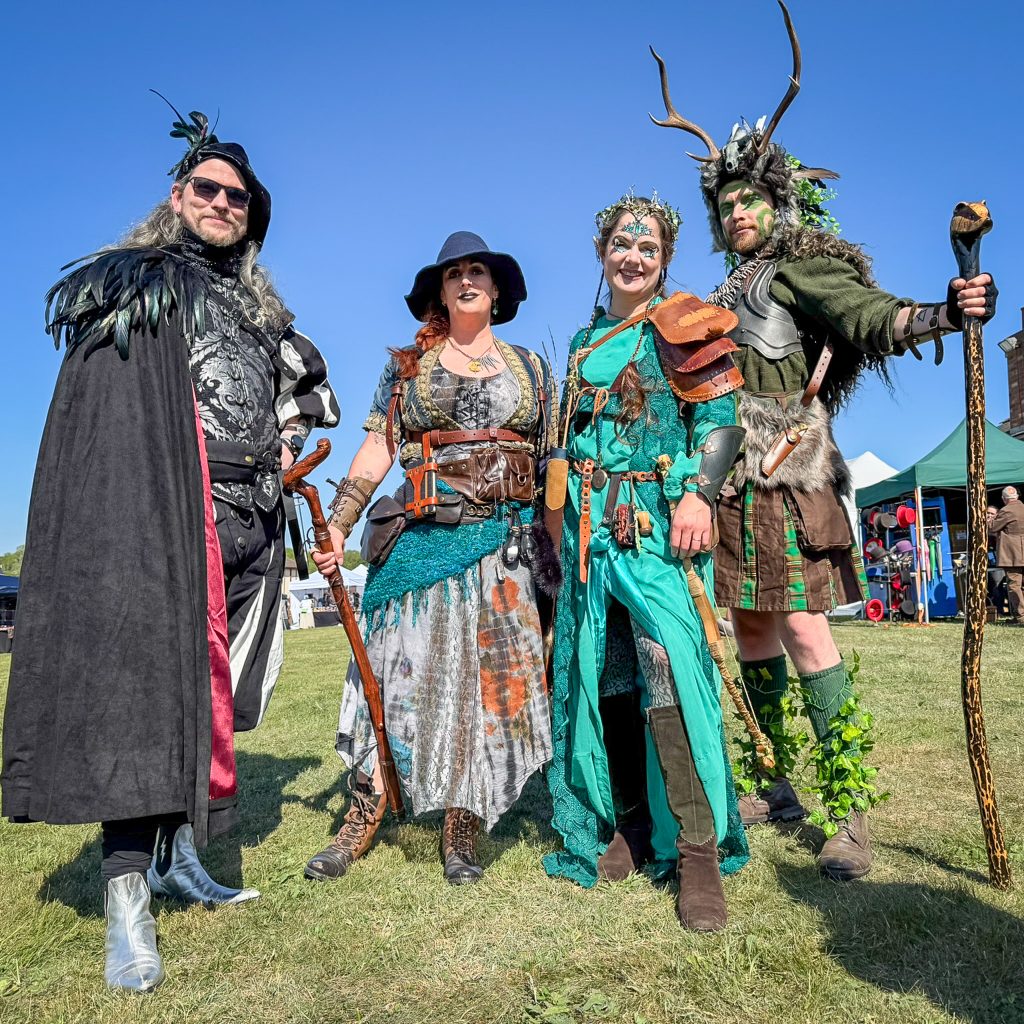 Four adults in elaborate fantasy costumes posing outdoors on a sunny day, wielding staffs and wearing woodland-themed attire.