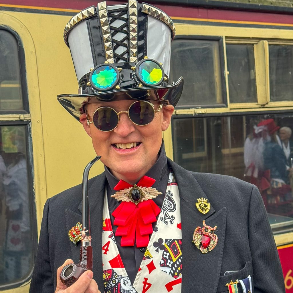 Person in a dark suit with a bright red bow tie, wearing a studded decorative top hat and iridescent round glasses, standing in front of a yellow train behind them.