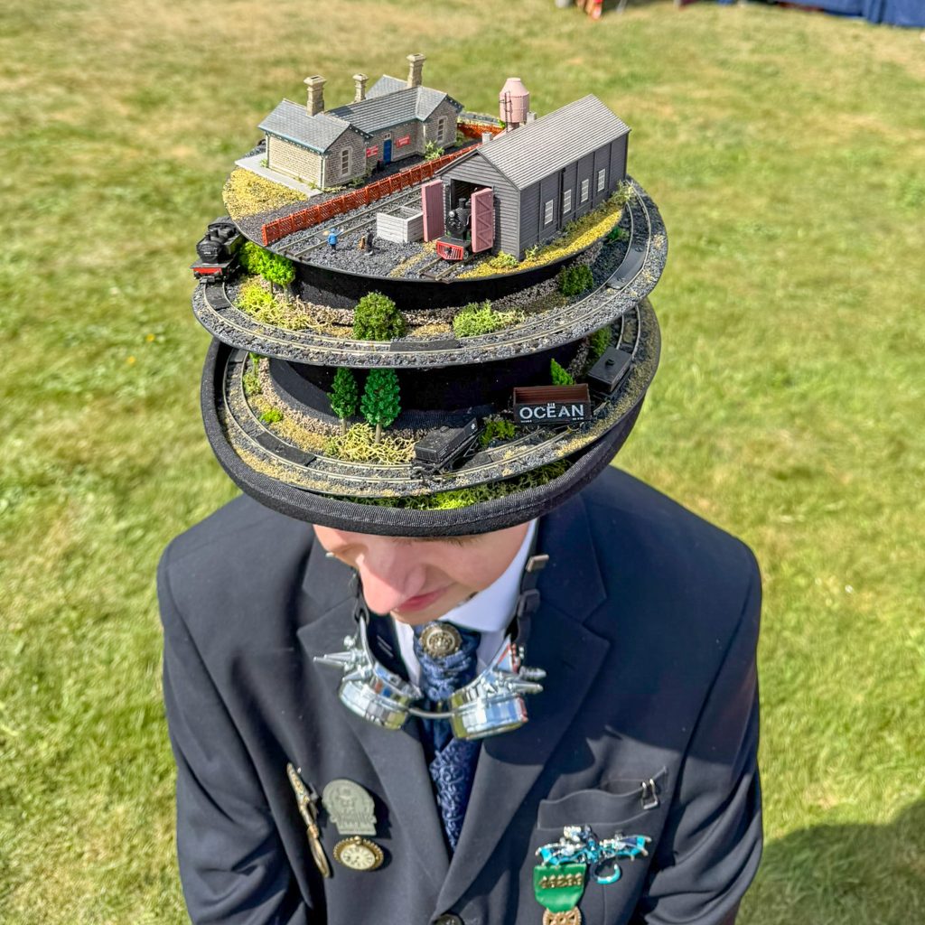 Man in a suit wearing a tall black hat that features a detailed miniature model train scene on top, with tracks, buildings, and tiny figures outdoors.