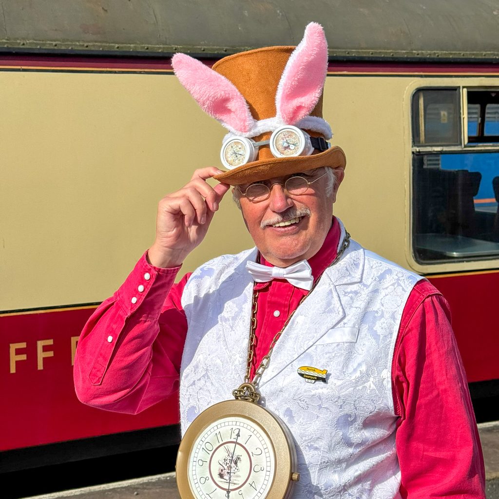 Person wearing a bunny-ear hat and steampunk-style goggles, outdoors with a large clock pendant on a chain at chest level.