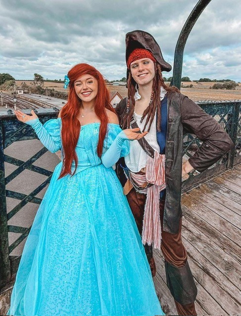 37ce039e-89ee-43e5-a2be-bc88b6fd5657 - Midland Railway - Butterley Two people in costumes posing on a wooden bridge: a woman in a blue princess dress with long red hair and a man in pirate attire with a hat.
