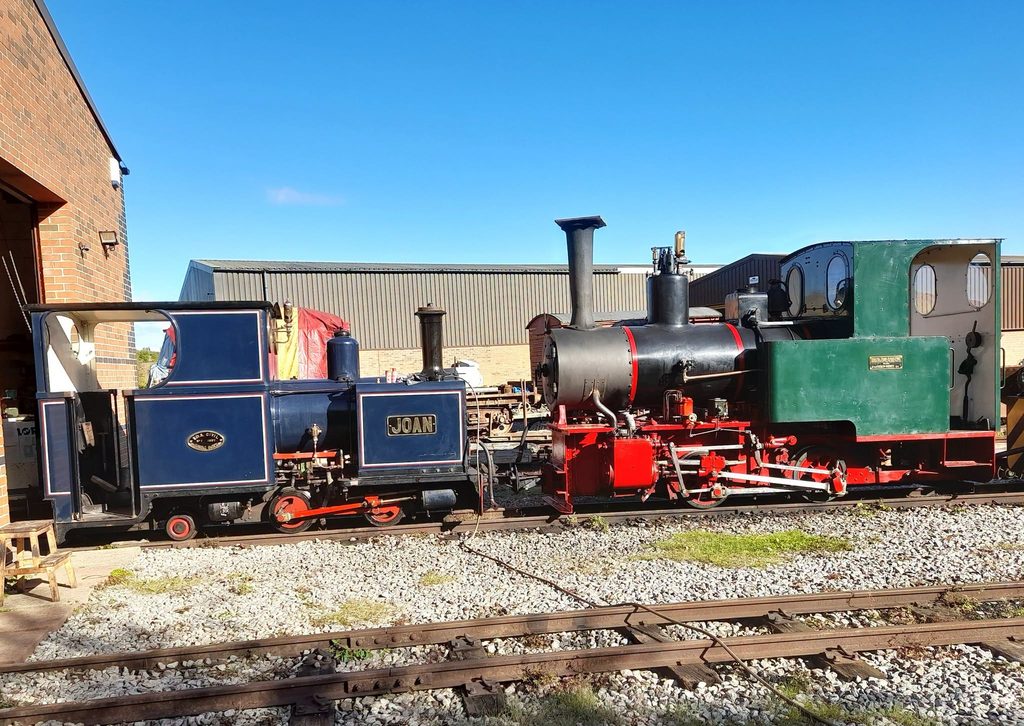 Historic steam locomotive on railroad tracks beside a brick building under a clear blue sky; blue cars and red wheels visible at the front with a green engine section on the right side of the image.