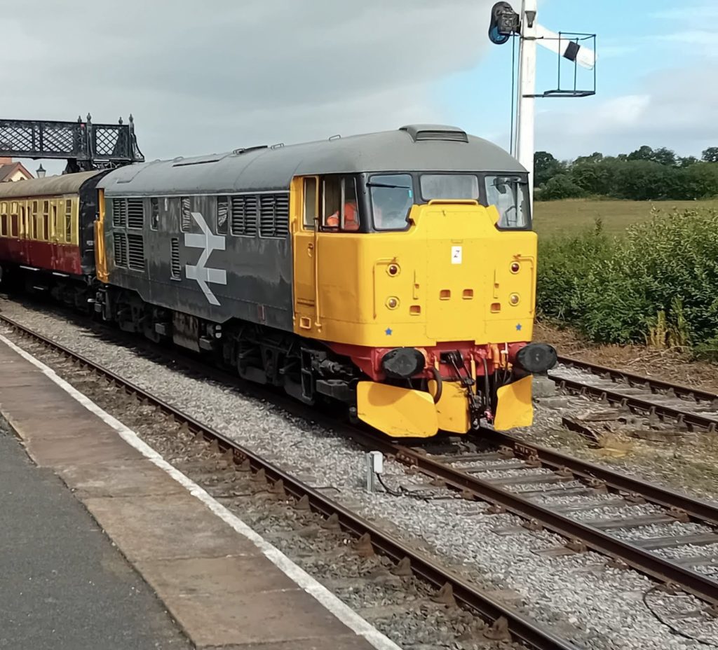 Diesel locomotive painted gray and yellow at a platform with passenger cars behind it on the tracks.