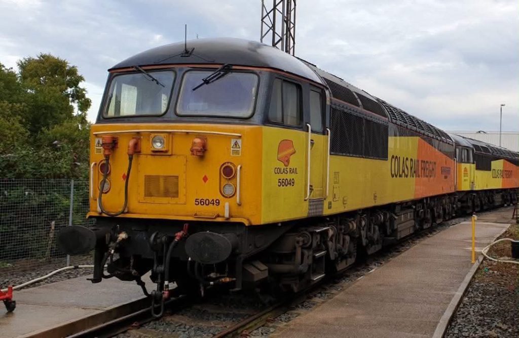 Front-and-side view of a Colas Rail Freight diesel locomotive in yellow and orange livery, numbered 56049, parked on a rail line with a trailing line of cars in the background.