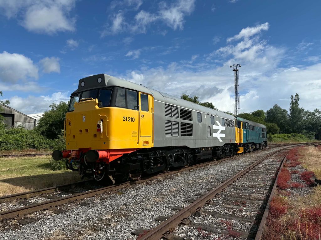 Diesel locomotive in yellow and gray on a railway track under a blue sky with white clouds, 31210 visible on the side at the front.