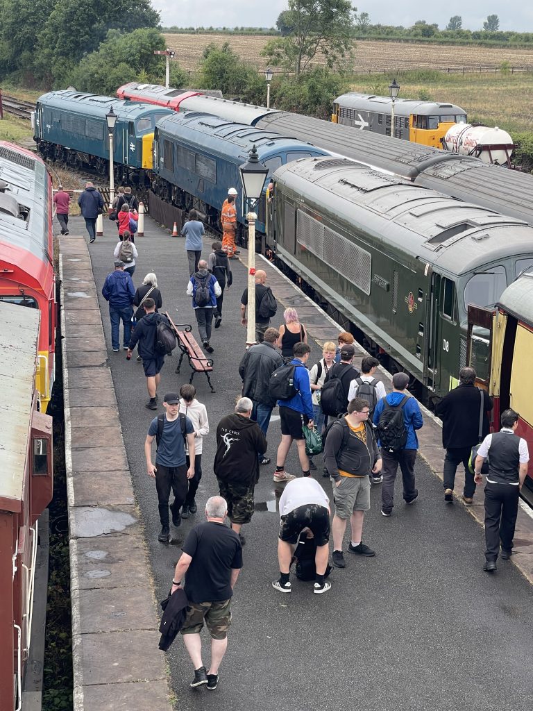 44 45 - Midland Railway - Butterley Crowded train platform with numerous people walking between blue and green locomotives on both sides, and fields in the background.