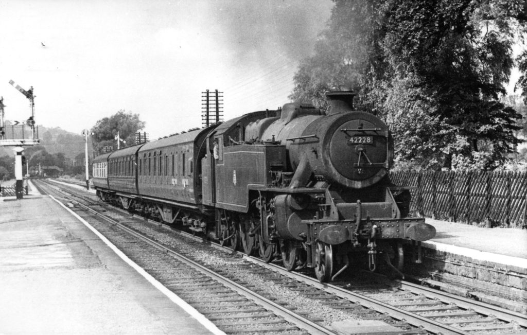 Fleming_P394 - Midland Railway - Butterley Black-and-white photo of a steam locomotive #42228 pulling passenger cars at a station platform with trees in the background.